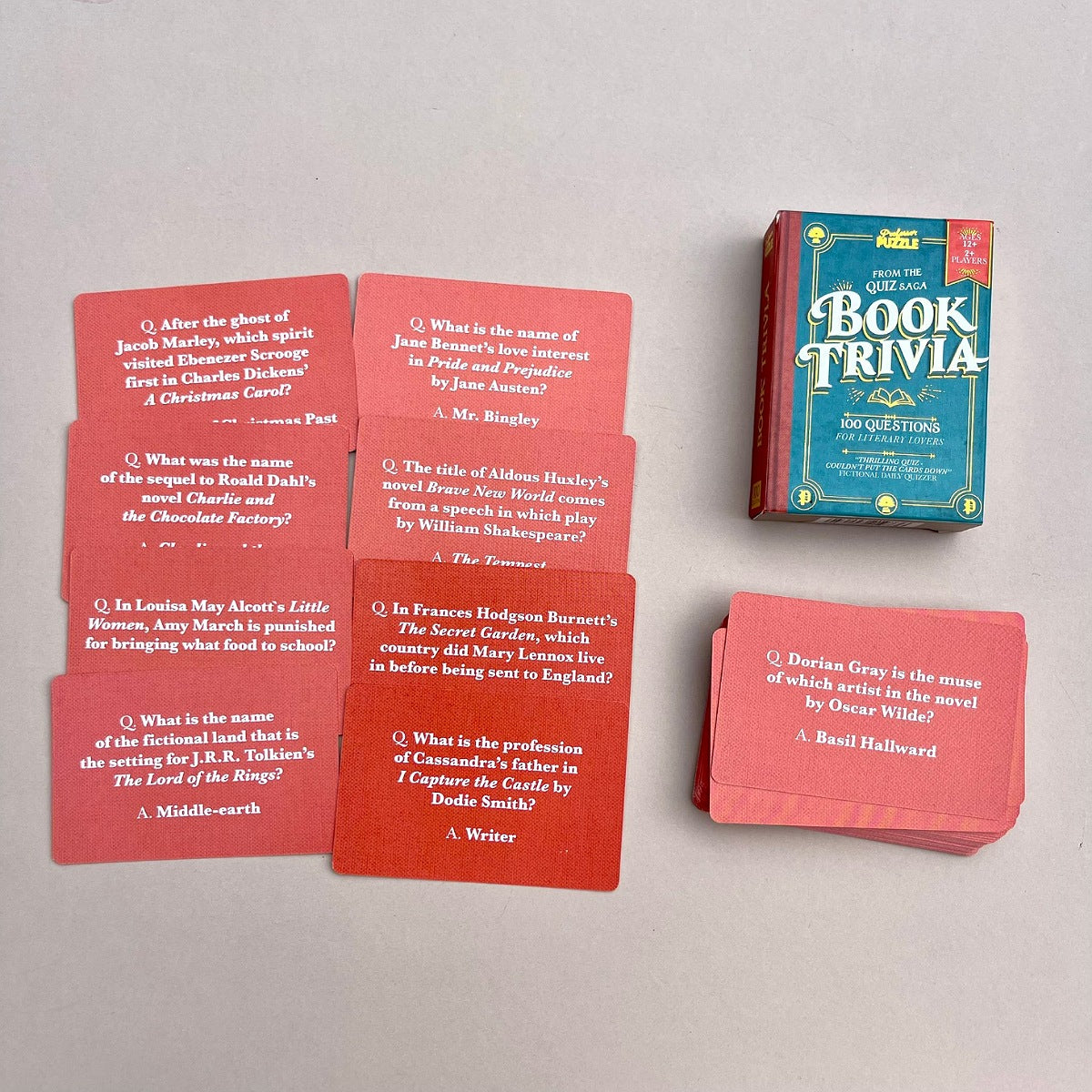A boxed deck of red trivia cards and the front cover of the deck are arranged on a light grey surface. The cards show multiple choice trivia questions, and the box is labeled 'The Book Trivia Deck'.