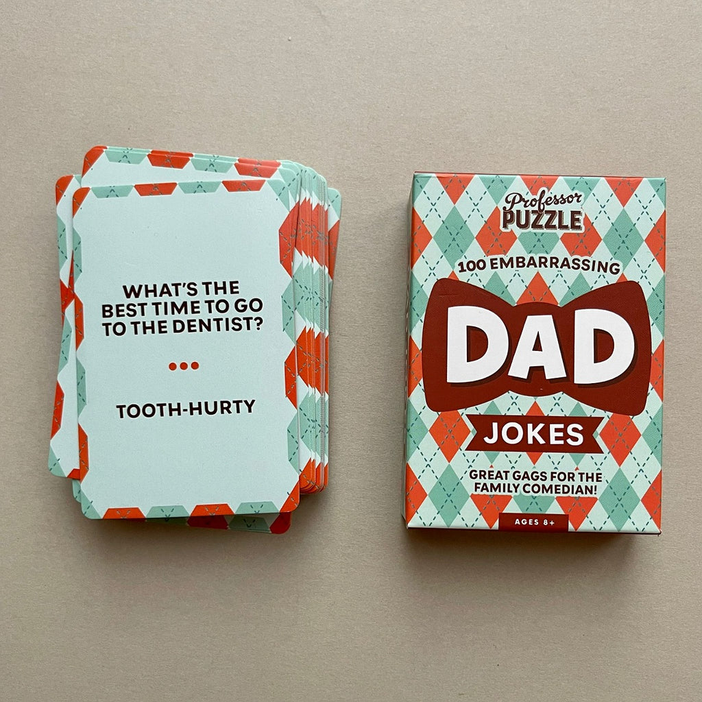 A box and a stack of cards on a plain beige surface. The box reads "Professor Puzzle" and "Dad Jokes," with a retro argyle pattern in mint green, coral, and white.