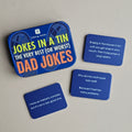 A set of four blue joke cards with white text, displayed on a light beige surface next to a matching blue tin box labeled 'Jokes in a Tin The Very Best (or Worst) Dad Jokes'.