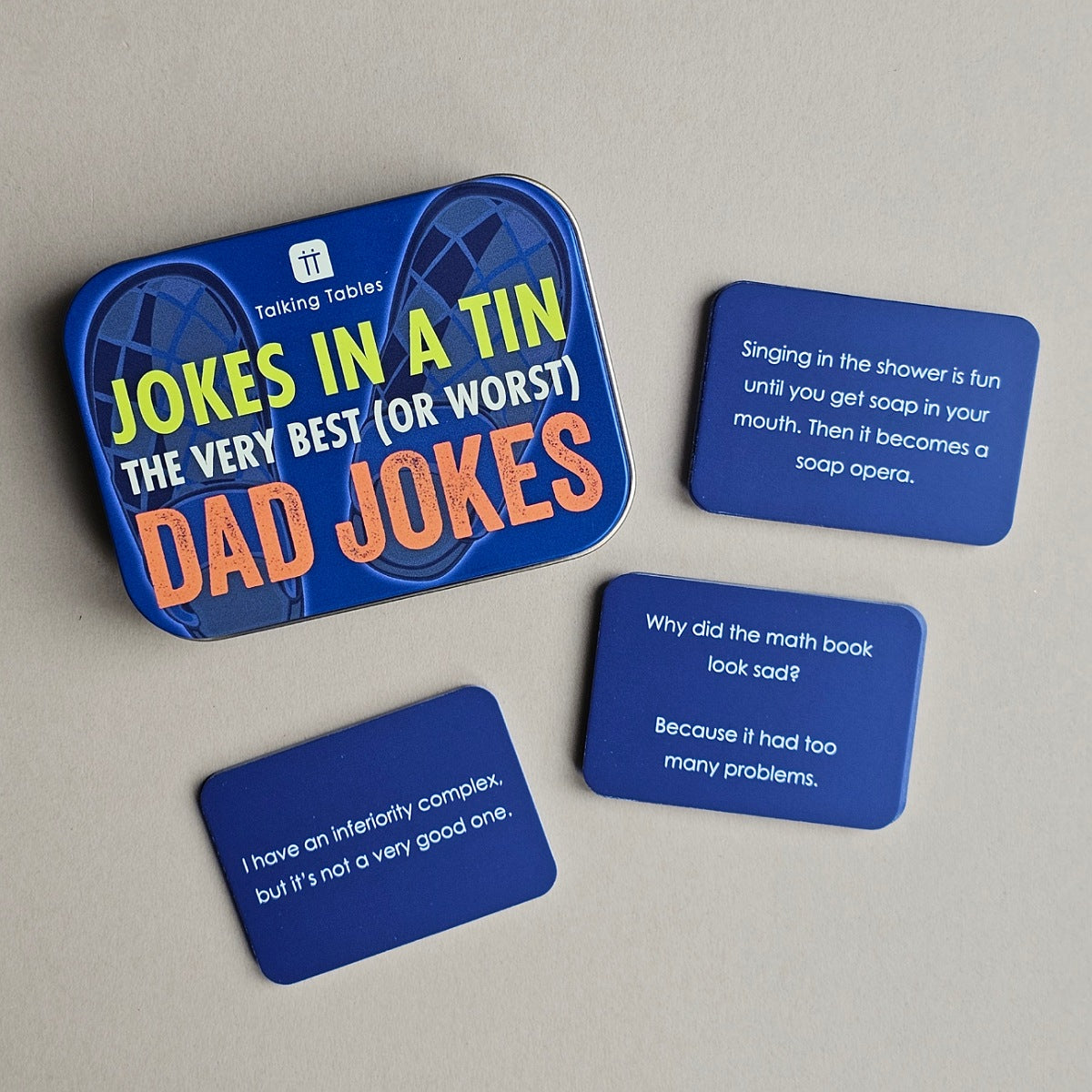 A set of four blue joke cards with white text, displayed on a light beige surface next to a matching blue tin box labeled 'Jokes in a Tin The Very Best (or Worst) Dad Jokes'.
