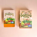 Two boxed card decks for Green Fingers Gardening Trivia, shown standing upright on a pale pink background. The left box is white with an orange base and colorful garden artwork; the right box is beige with matching artwork and text reading '100 Gardening Trivia Questions.