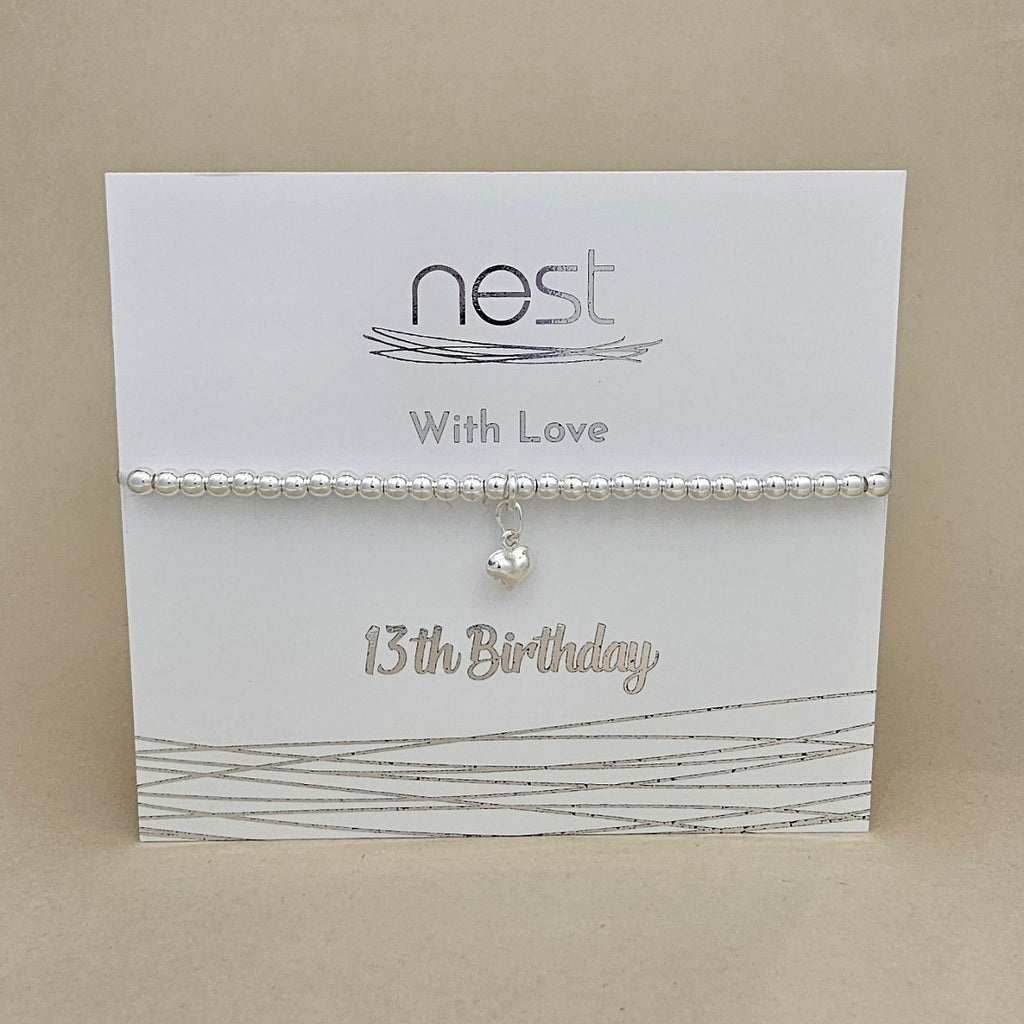A silver beaded birthday bracelet with a small dangling heart charm, displayed on a white card that reads 'nest With Love 13th Birthday' against a beige background.