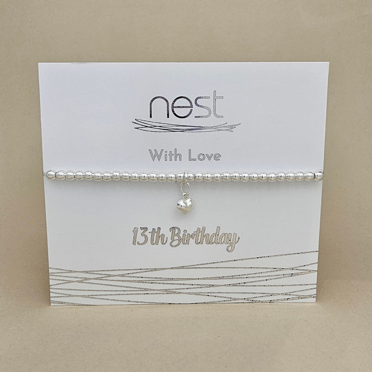 A silver beaded birthday bracelet with a small dangling heart charm, displayed on a white card that reads 'nest With Love 13th Birthday' against a beige background.