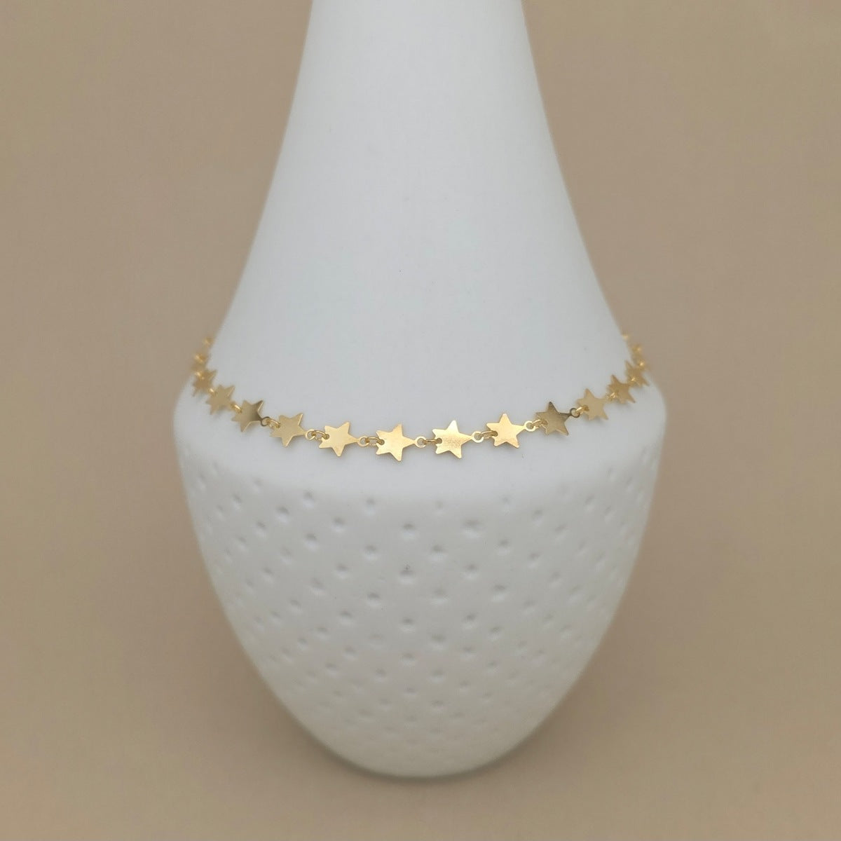 A delicate gold star chain anklet displayed on a white textured vase against a solid beige background.