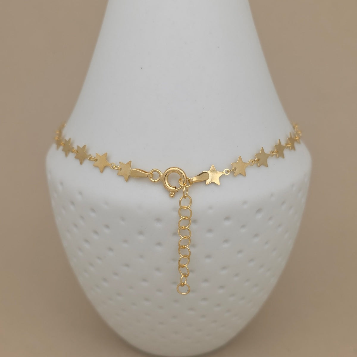 A gold star charm anklet displayed on a white textured jewelry bust against a solid beige background.