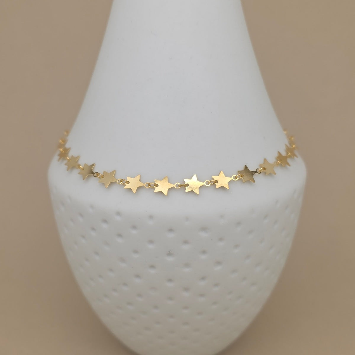A delicate gold star charm anklet displayed on a white textured cone jewelry stand against a warm beige background.