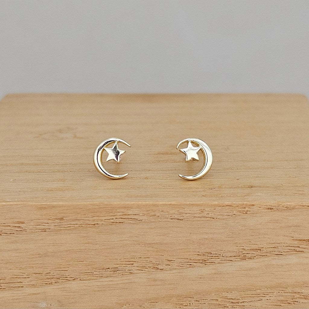 A pair of small gold crescent moon and star stud earrings displayed on a light wood surface against a neutral gray background.