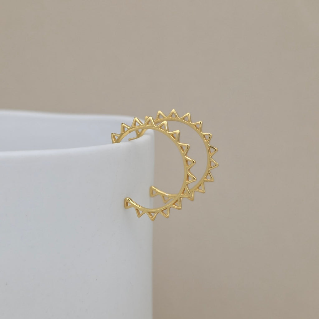 A gold hoop earrings with triangular sunburst details on the ends, displayed on a white cylindrical stand against a neutral beige background.