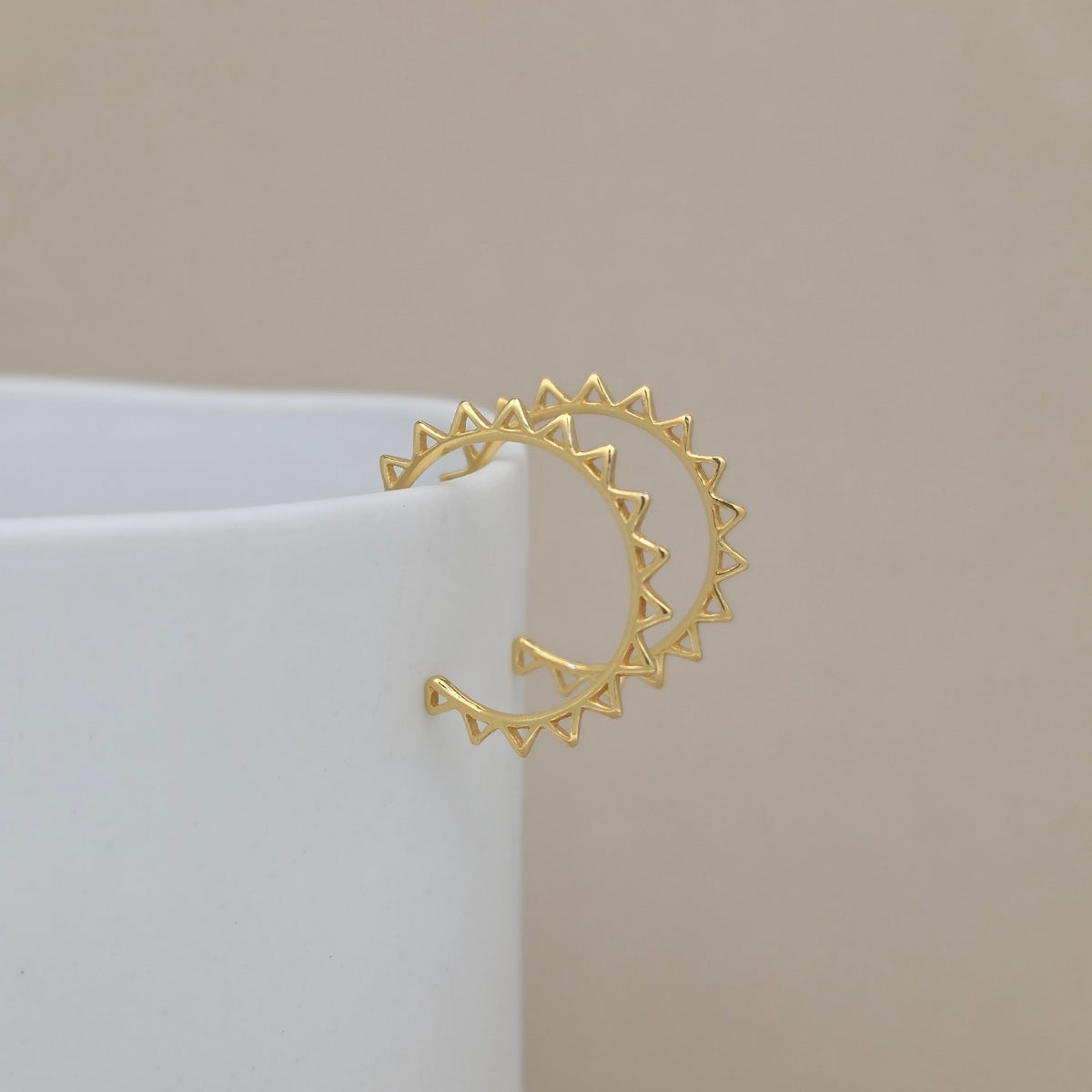 A gold hoop earrings with triangular sunburst details on the ends, displayed on a white cylindrical stand against a neutral beige background.