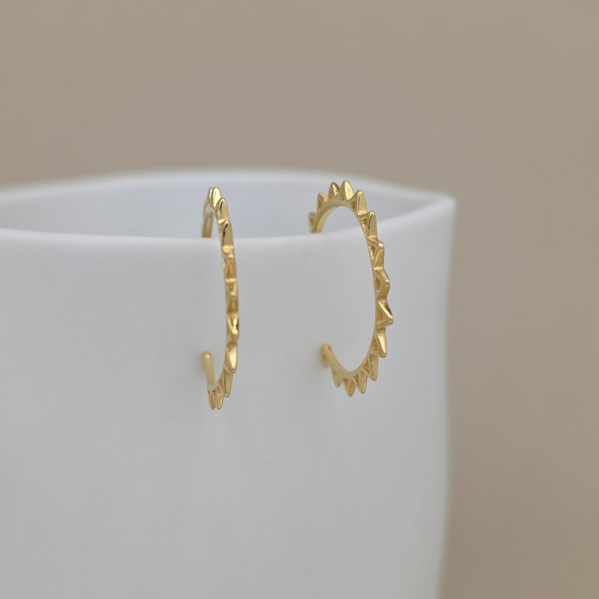 A pair of delicate gold hoop earrings with a textured, scalloped edge, displayed on a white ceramic cup against a soft beige background.
