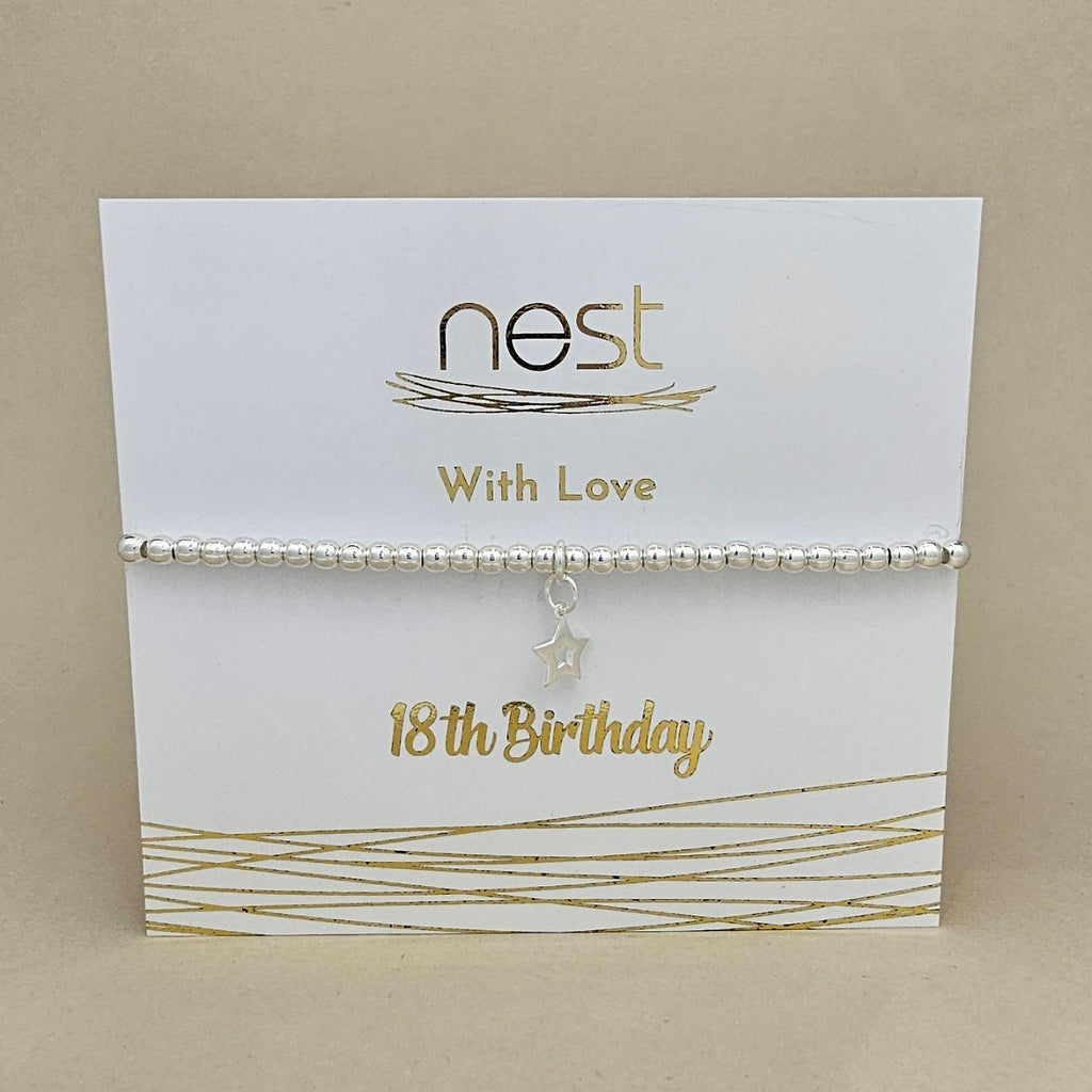A silver beaded bracelet with a dangling star charm, presented on a white card with gold foil text reading 'nest With Love 18th Birthday' against a neutral beige background.