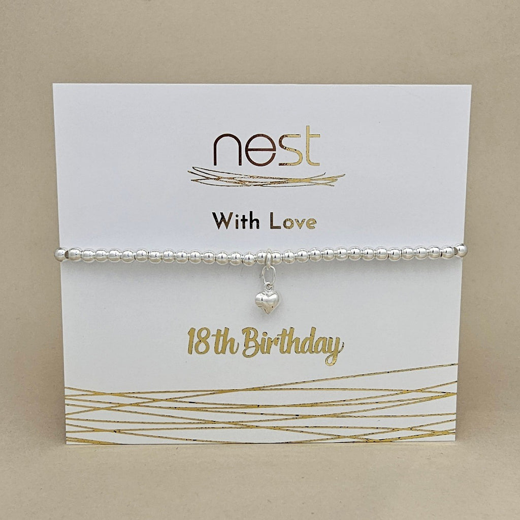 A silver beaded bracelet with a small dangling heart charm, displayed on a white card that reads 'nest With Love 18th Birthday' against a beige background.