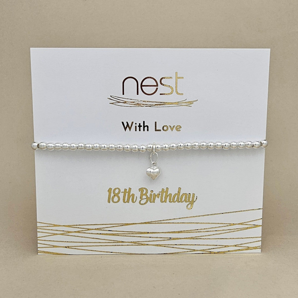 A silver beaded bracelet with a small dangling heart charm, displayed on a white card that reads 'nest With Love 18th Birthday' against a beige background.