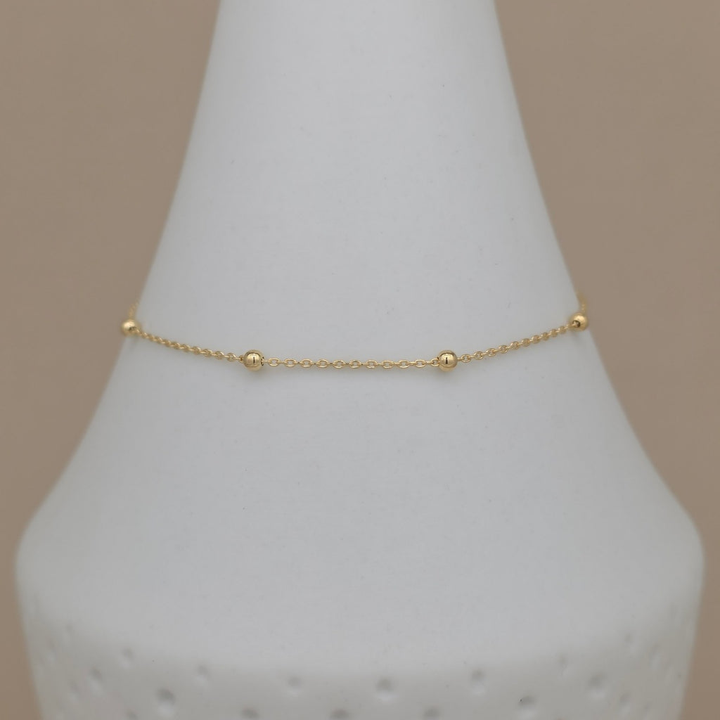 A delicate gold chain bracelet with three small bead accents, displayed on a white cone jewelry stand against a solid beige background.