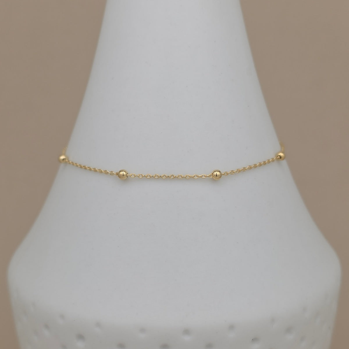A delicate gold chain bracelet with three small bead accents, displayed on a white cone jewelry stand against a solid beige background.
