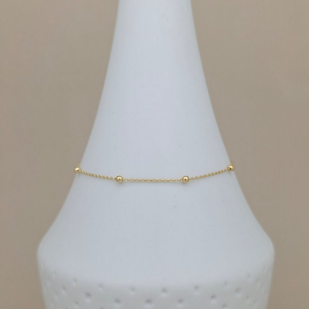 A delicate gold chain bracelet with small bead accents, displayed on a white cone jewelry stand against a soft beige background.