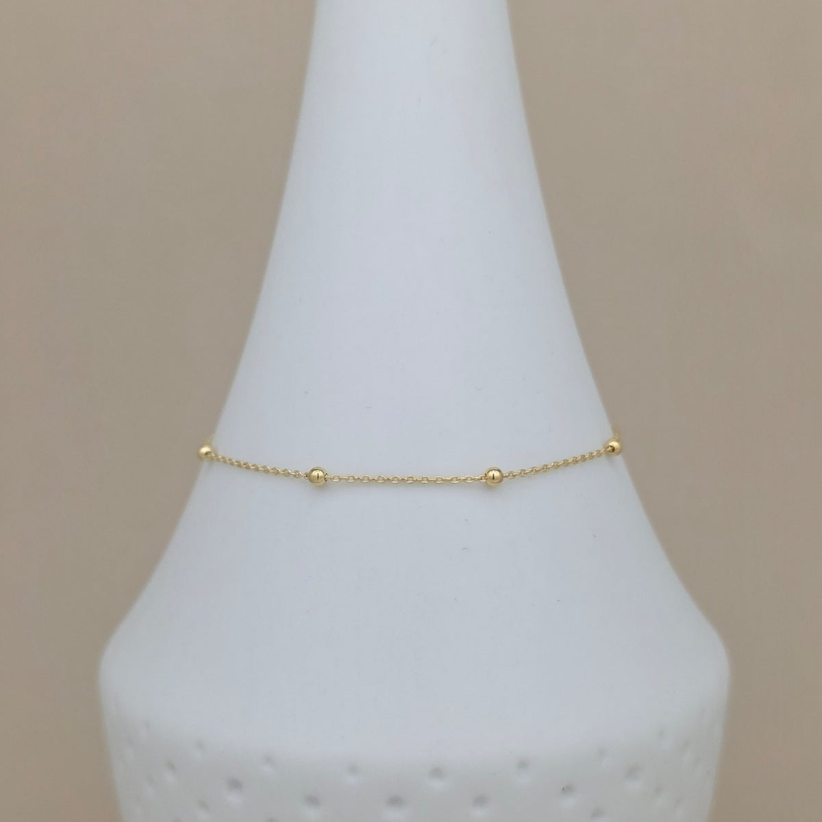 A delicate gold chain bracelet with small bead accents, displayed on a white cone jewelry stand against a soft beige background.