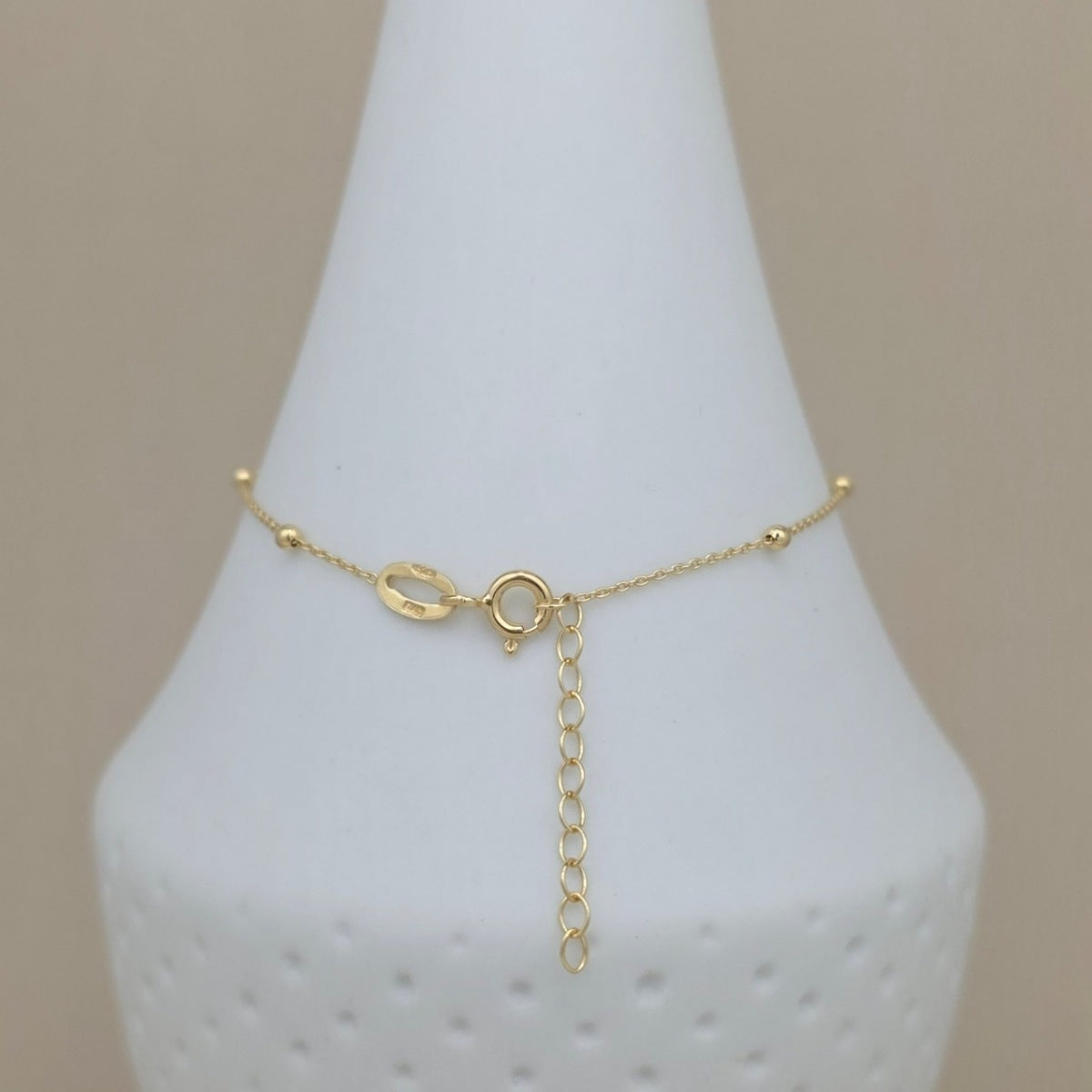 A delicate gold chain anklet with a unique open-link charm and an adjustable drop chain extender, displayed on a white cone-shaped jewelry stand against a neutral beige background.