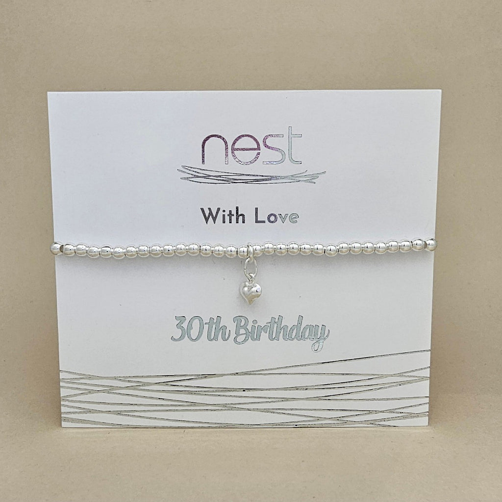 A silver beaded bracelet with a small dangling heart charm, displayed on a white gift card that reads 'nest With Love 30th Birthday' against a beige background.
