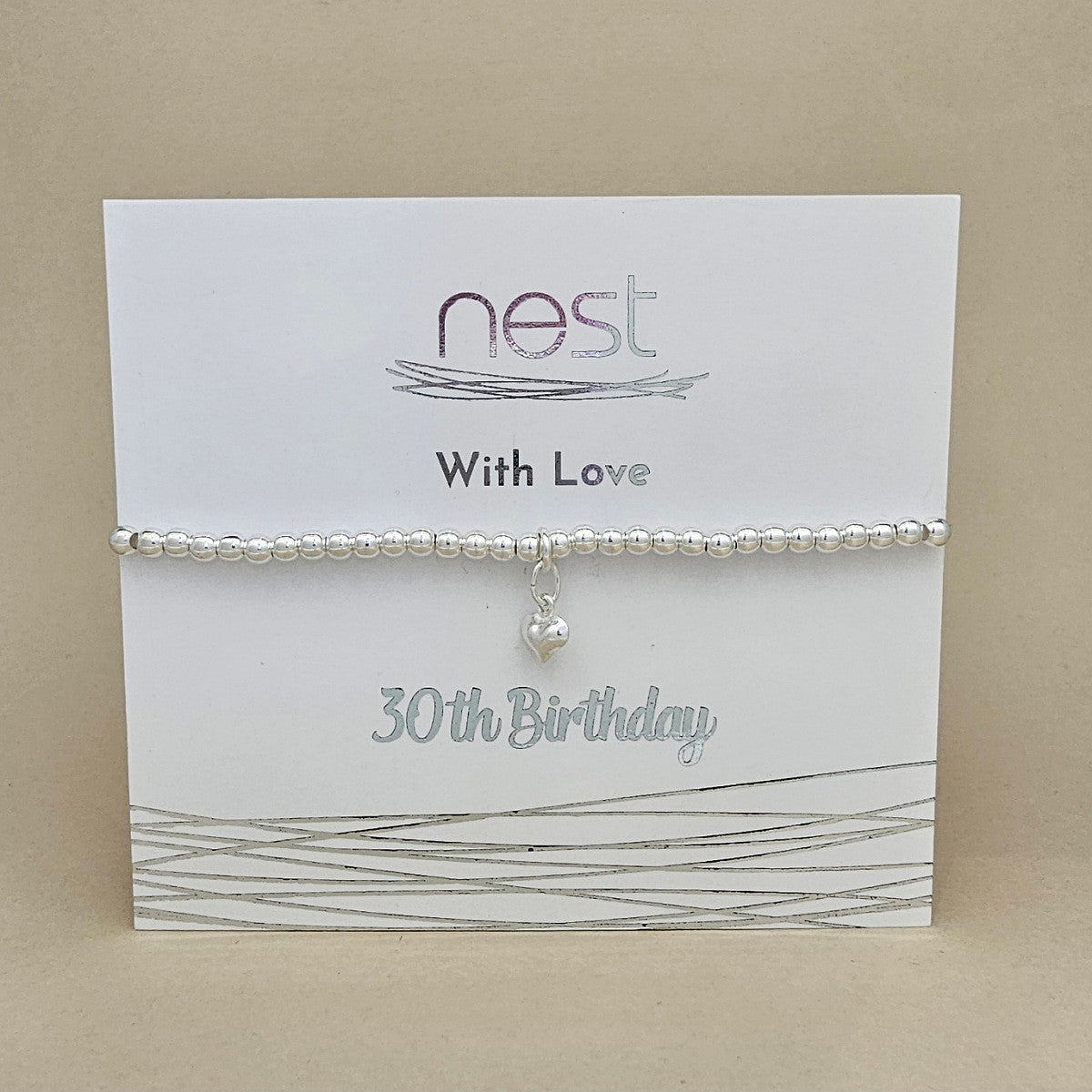 A silver beaded bracelet with a small dangling heart charm, displayed on a white gift card that reads 'nest With Love 30th Birthday' against a beige background.