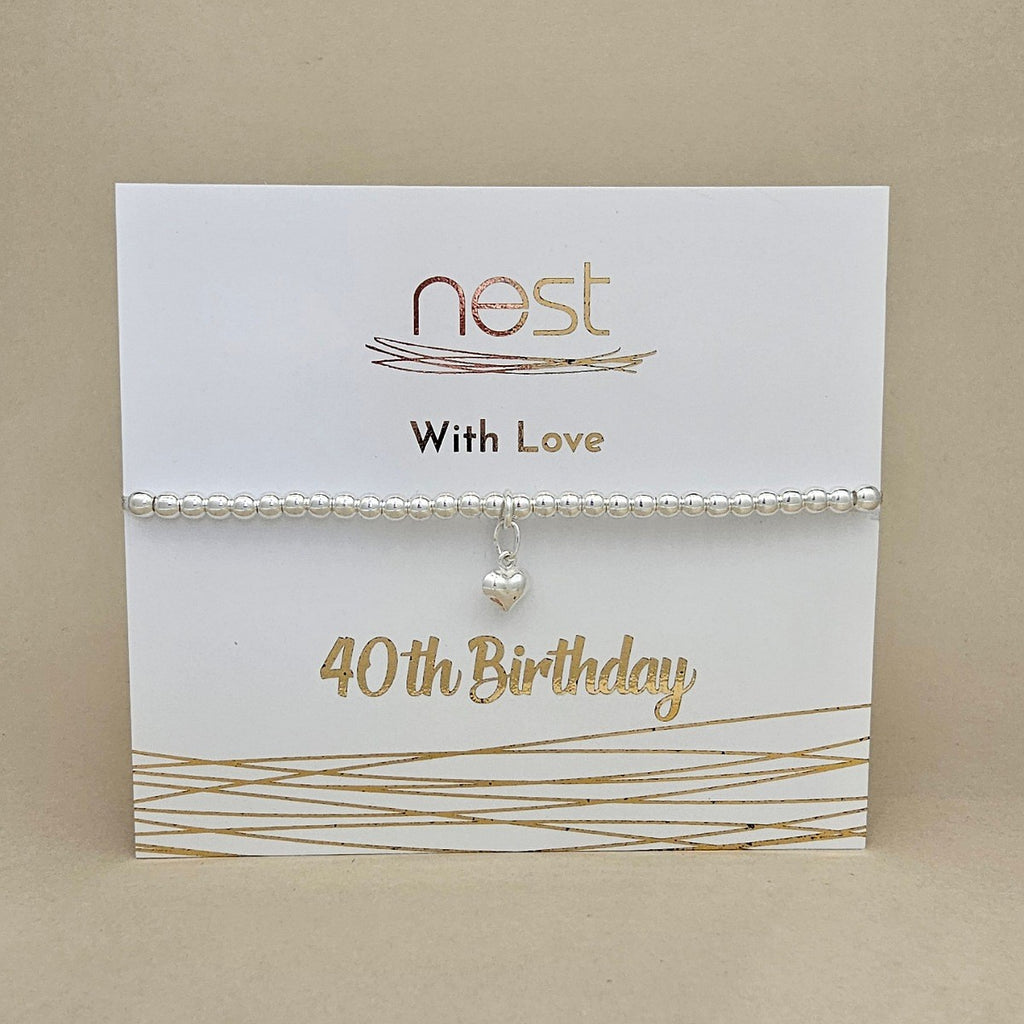 A silver beaded bracelet with a small heart charm, displayed on a white card printed with 'nest With Love 40th Birthday' in gold lettering, set against a neutral beige background.