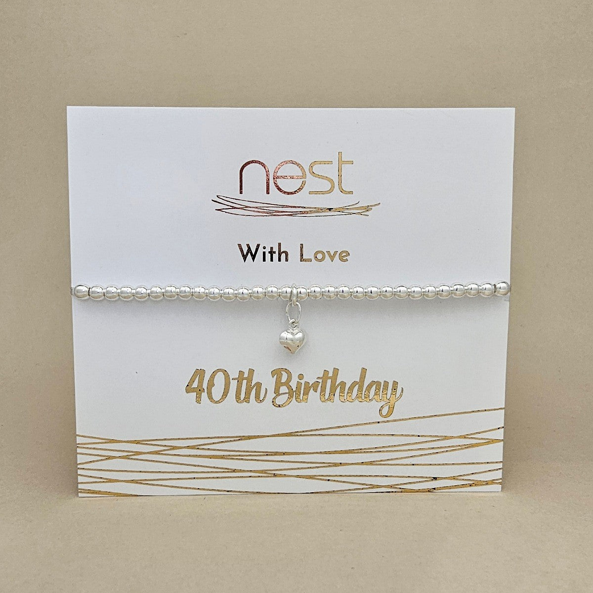 A silver beaded bracelet with a small heart charm, displayed on a white card printed with 'nest With Love 40th Birthday' in gold lettering, set against a neutral beige background.
