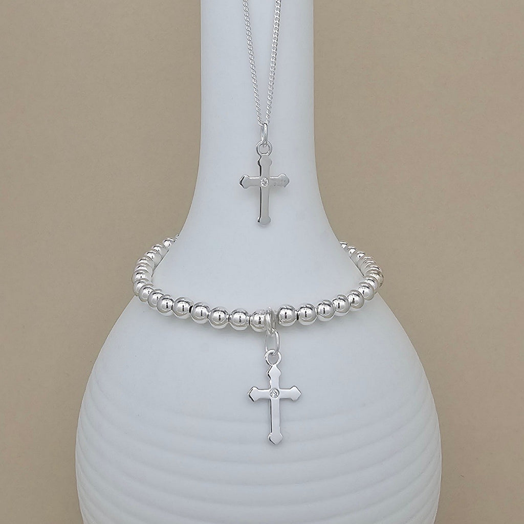 A silver beaded bracelet and matching necklace with a cross pendant, displayed on a white ribbed vase against a beige background.