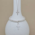 A silver beaded bracelet and matching necklace with a cross pendant, displayed on a white ribbed vase against a beige background.