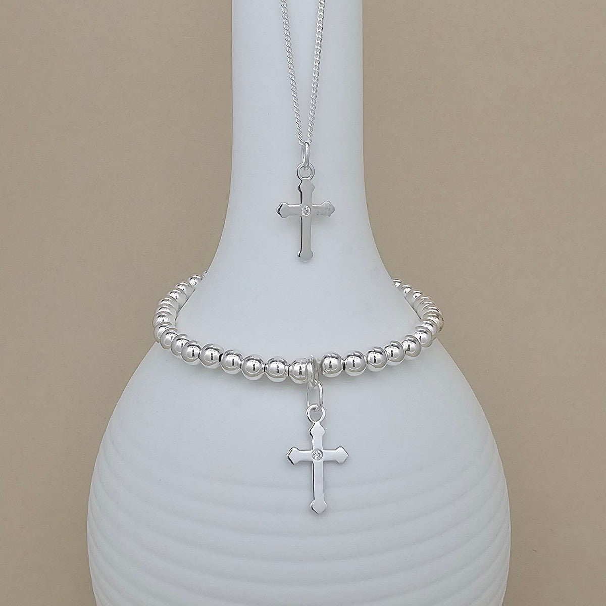 A silver beaded bracelet and matching necklace with a cross pendant, displayed on a white ribbed vase against a beige background.