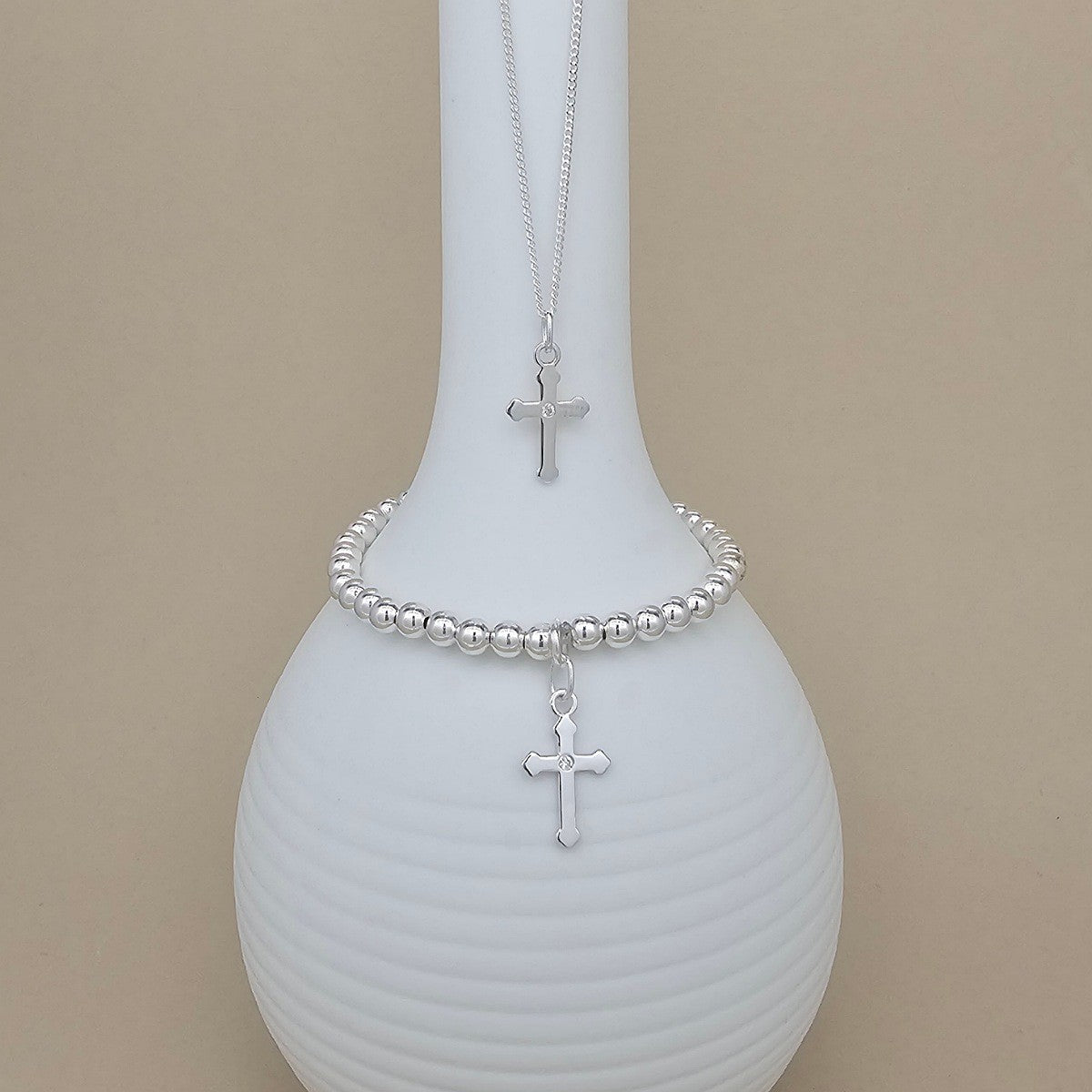 A silver beaded bracelet with a cross pendant is displayed on a white ribbed vase, with a matching silver necklace hanging above it against a beige background.