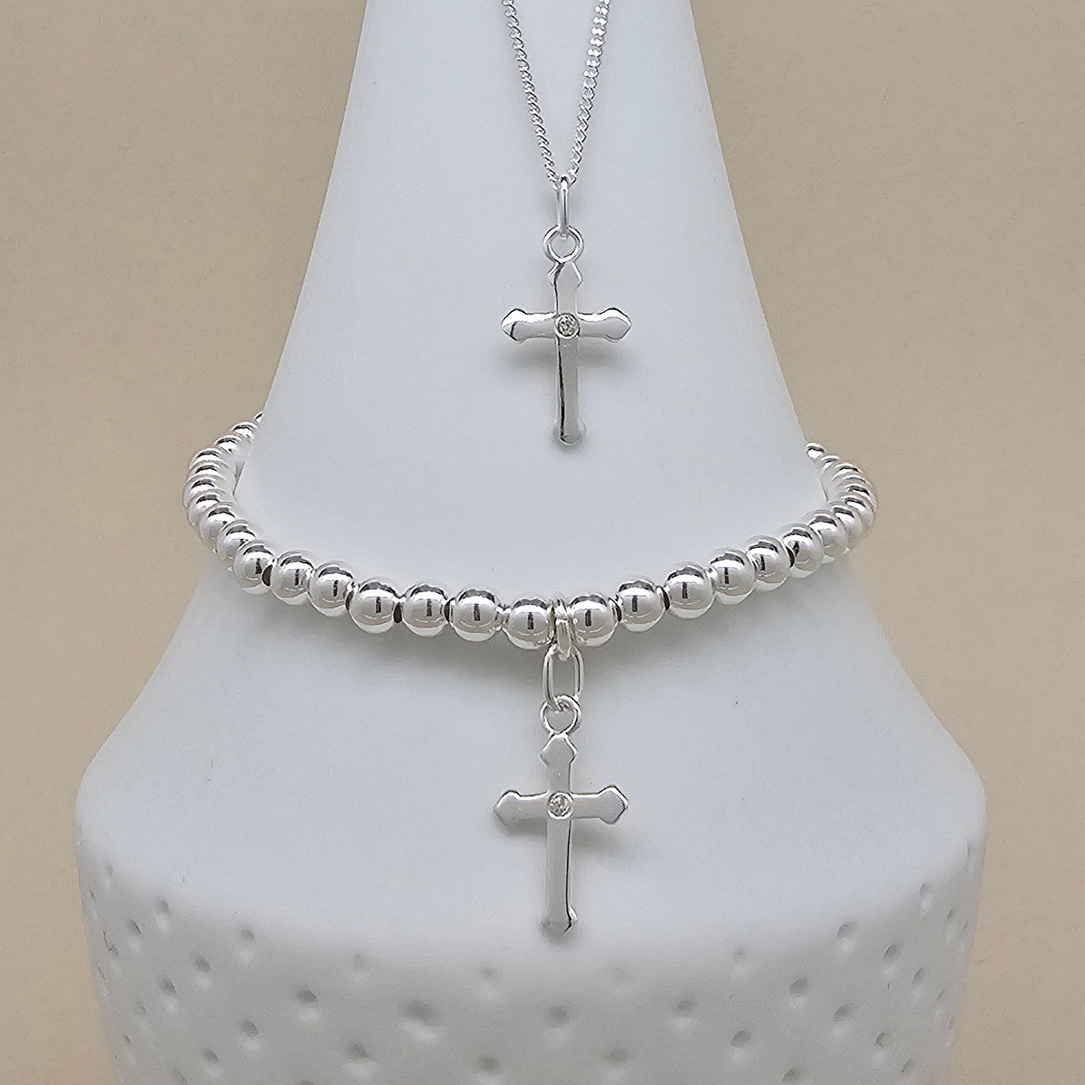 A silver cross necklace and matching beaded bracelet displayed on a white cone-shaped jewelry stand against a light beige background.