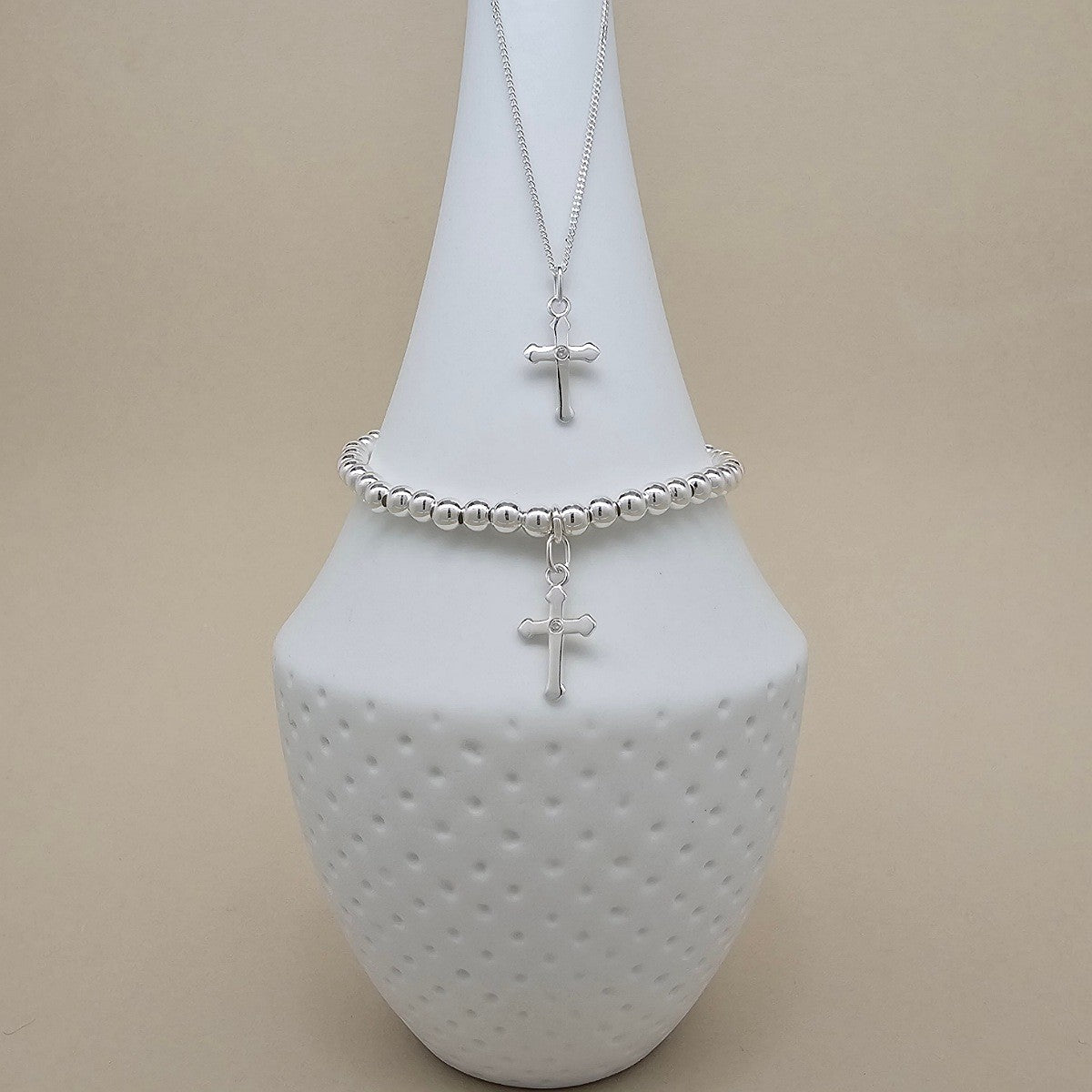 A silver necklace and matching beaded bracelet with small cross pendants, displayed on a white ceramic vase against a warm beige background.