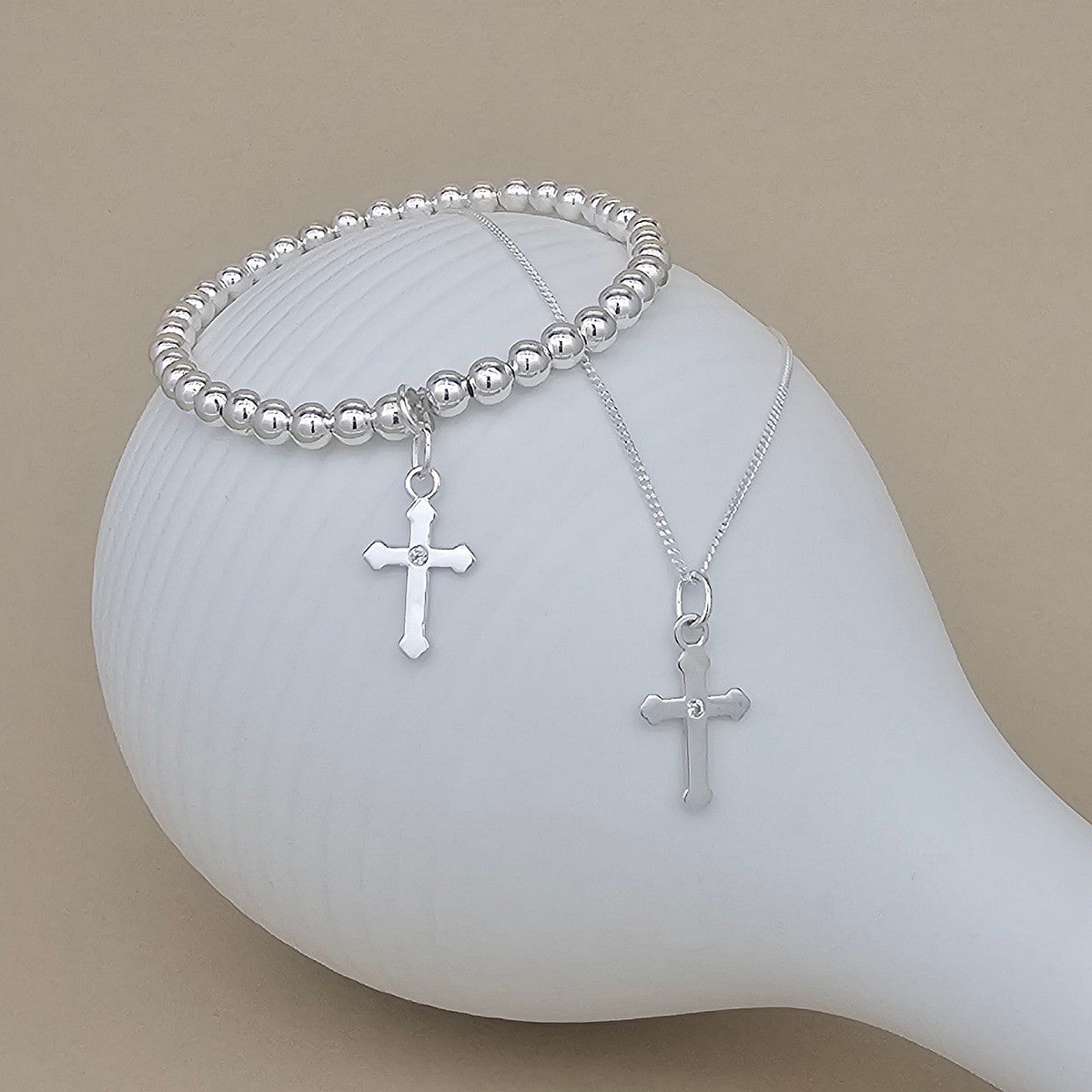 A silver beaded bracelet and a silver cross pendant necklace displayed on a white ceramic mannequin against a beige background.