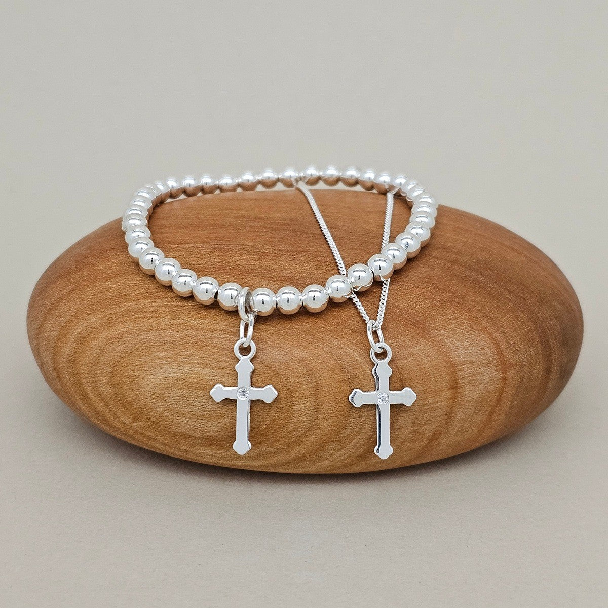 A silver bracelet and silver necklace with silver cross charms on a wooden rock.