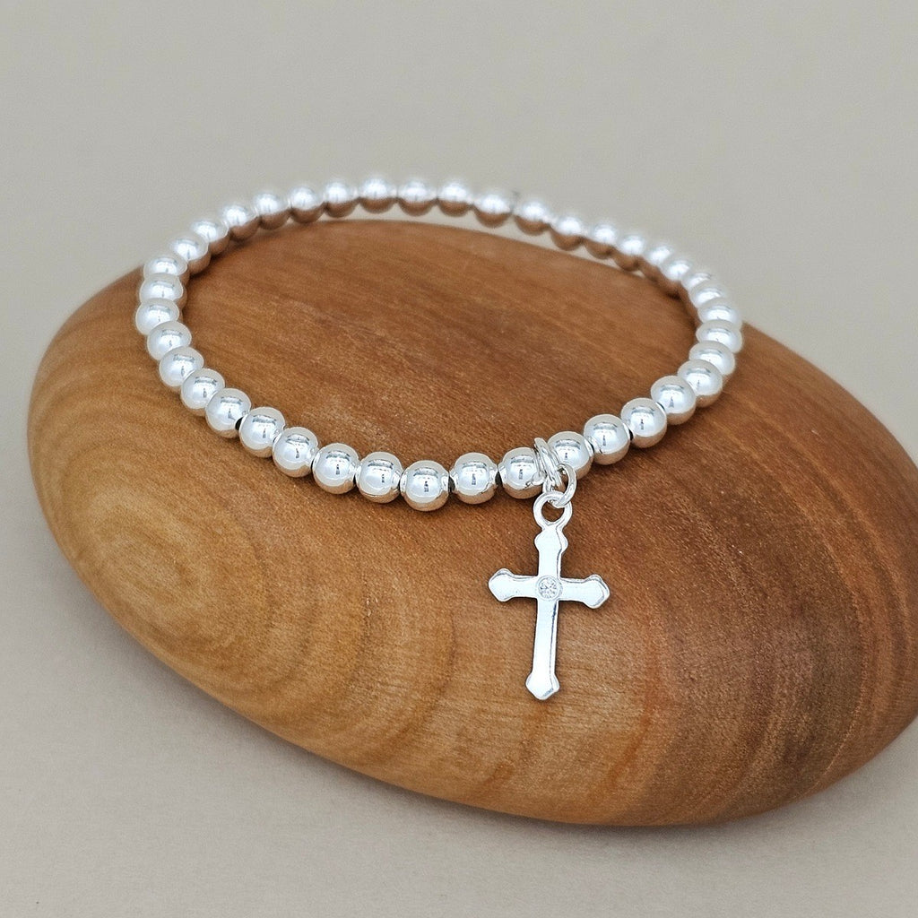 A silver beaded bracelet with a small cross pendant, displayed on a smooth brown stone against a neutral beige background.