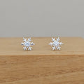 A pair of silver snowflake stud earrings with clear central gemstones, displayed on a light brown wooden block against a neutral gray background.
