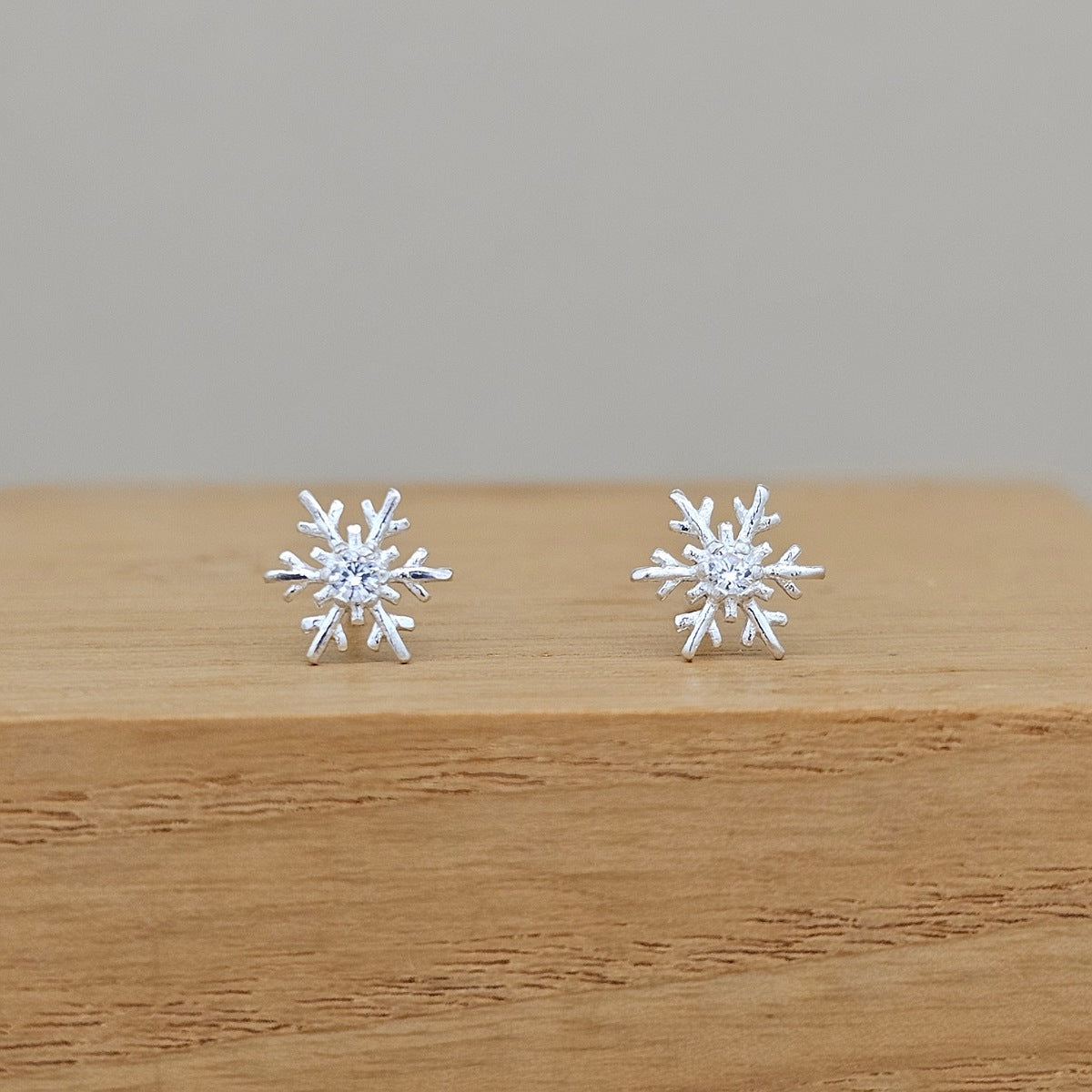 A pair of silver snowflake stud earrings with clear central gemstones, displayed on a light brown wooden block against a neutral gray background.