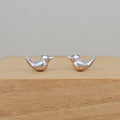 A pair of small silver bird stud earrings shaped like robins, displayed on a light wood block against a neutral gray background.