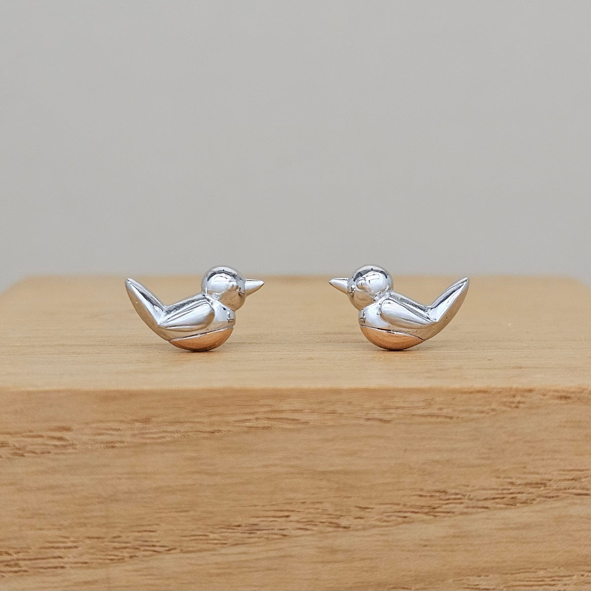 A pair of small silver bird stud earrings shaped like robins, displayed on a light wood block against a neutral gray background.
