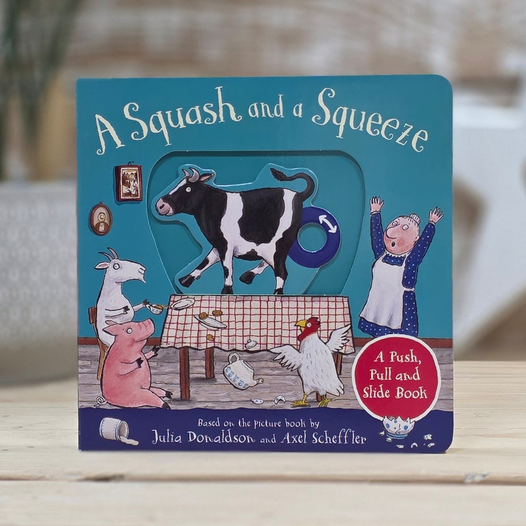 A children's push-pull and slide book titled "A Squash and a Squeeze" standing upright on a light wooden surface. The cover features colorful cartoon farm animals including a cow, pig, goat, chicken, and a woman around a table, with a teal background and a red circular badge that reads "A Push, Pull and Slide Book.