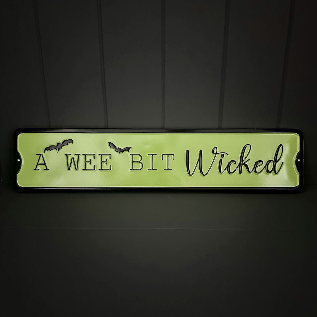 A rectangular novelty sign with a green background and black border, featuring the phrase 'A WEE BIT Wicked' in black lettering with bat silhouettes, shown against a dark panelled background.