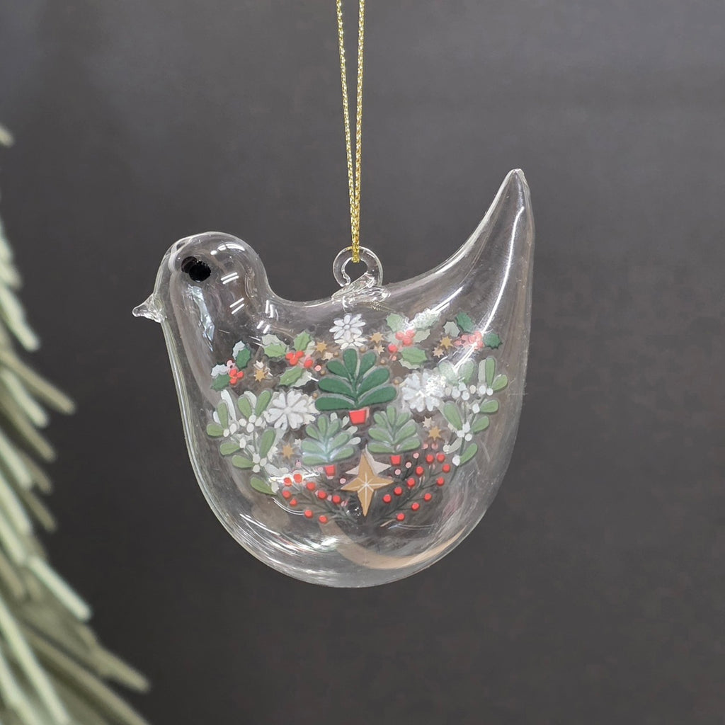 A clear glass bird Christmas ornament with a hand-painted winter floral design inside, hanging from a gold string against a dark grey background.