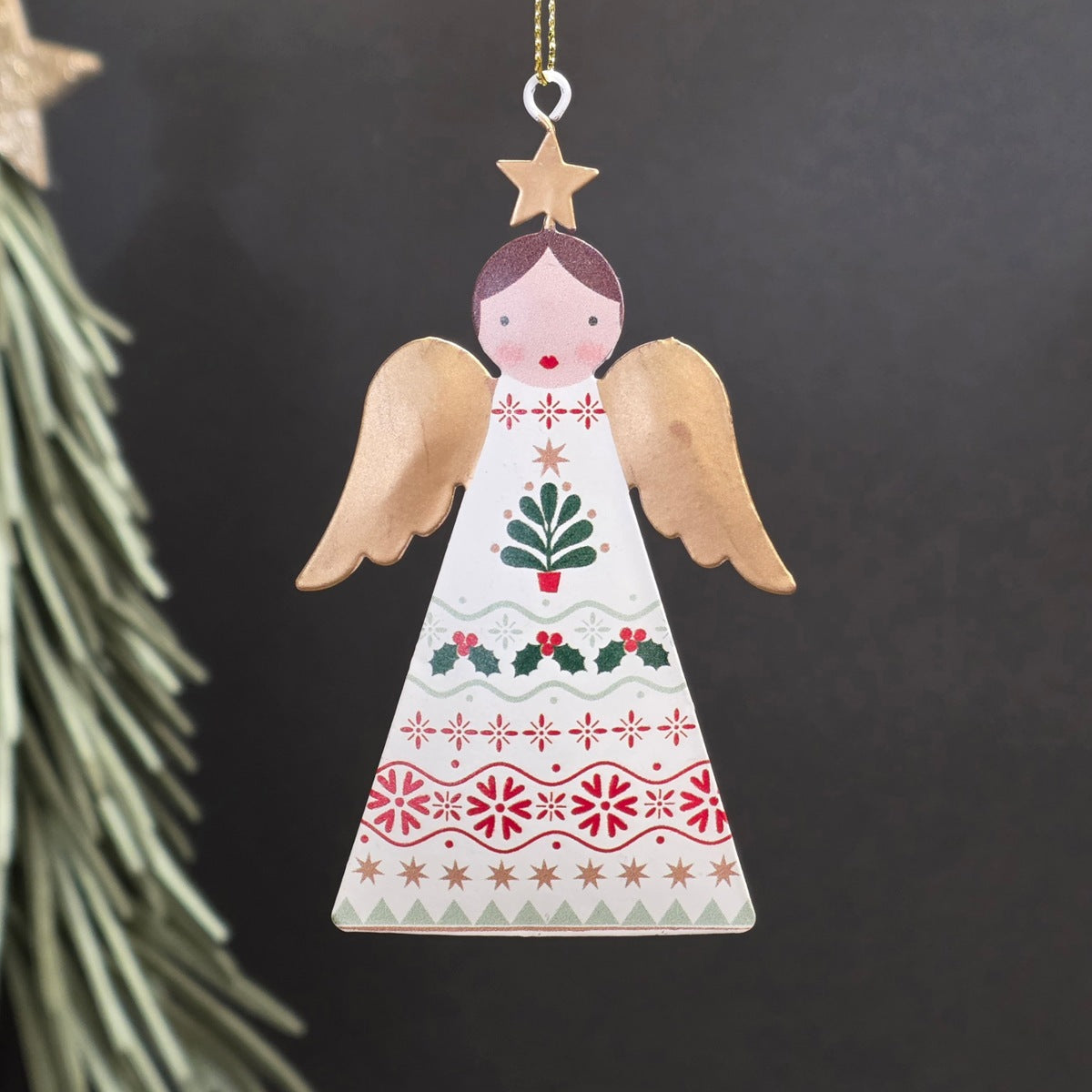 A decorative metal Christmas ornament shaped like an angel, featuring a cream-coloured body with hand-painted red, green, and gold holiday motifs and wings, hanging from a gold string against a dark background.