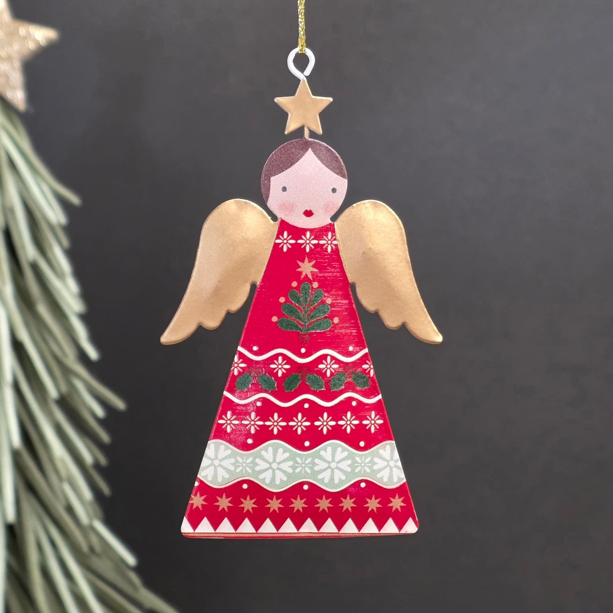 A decorative metal Christmas ornament shaped like an angel, featuring a red-coloured body with hand-painted red, green, and gold holiday motifs and wings, hanging from a gold string against a dark background.