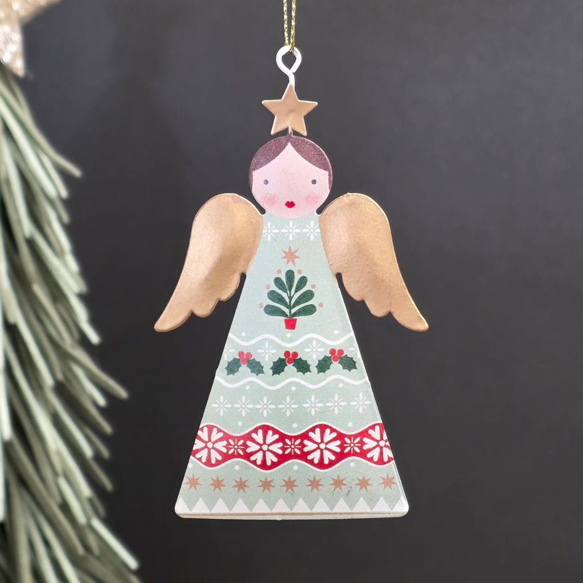 A decorative metal Christmas ornament shaped like an angel, featuring a light sage green-coloured body with hand-painted red, green, and gold holiday motifs and wings, hanging from a gold string against a dark background.