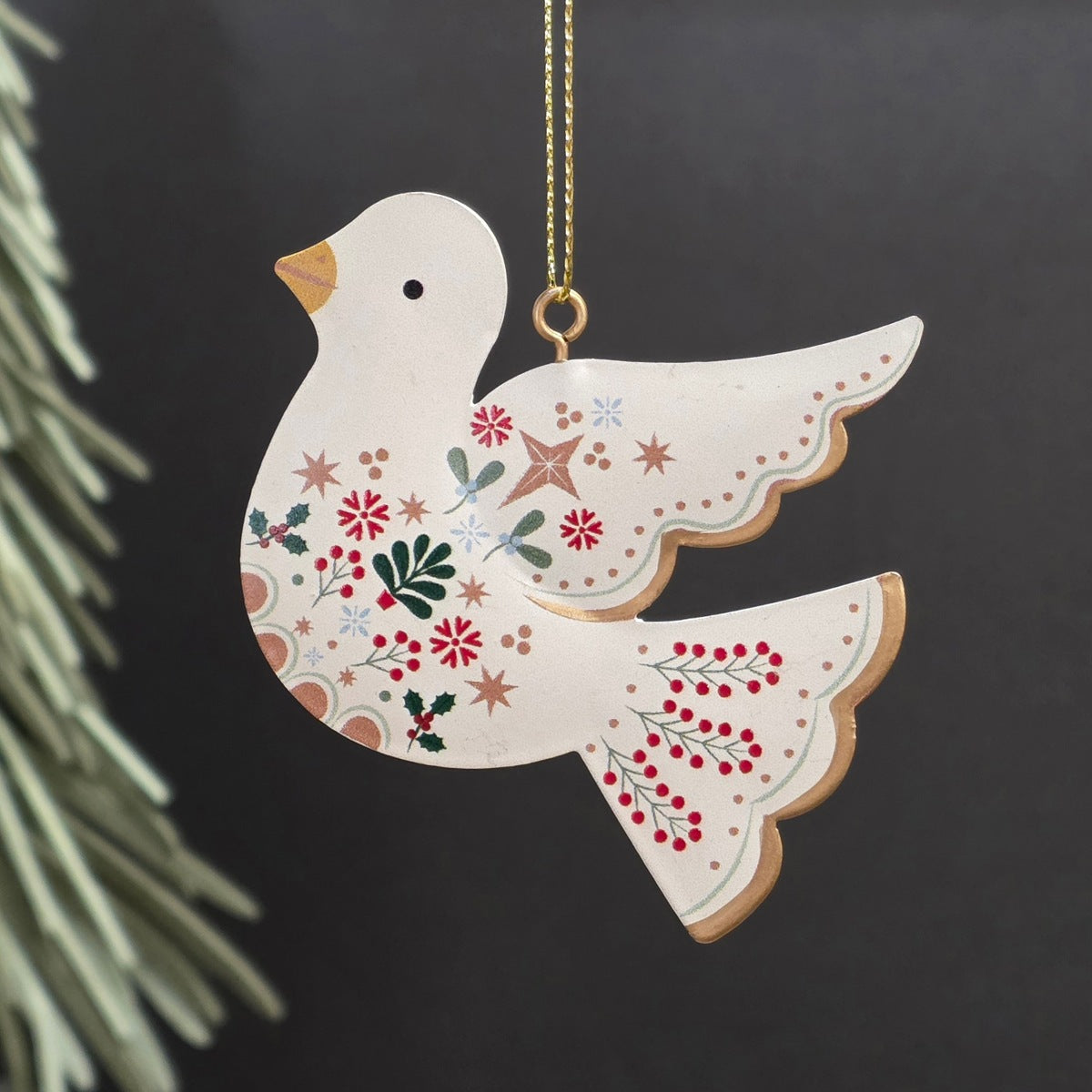 A decorative metal Christmas ornament shaped like a dove, featuring a cream-colored body with hand-painted red, green, and gold holiday motifs, hanging from a gold chain against a dark background.