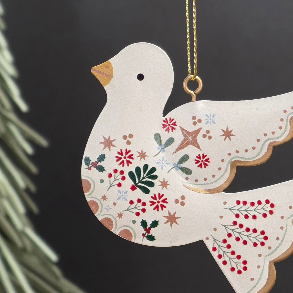 A decorative metal Christmas ornament shaped like a dove, featuring a cream-colored body with hand-painted red, green, and gold holiday motifs, hanging from a gold chain against a dark background.