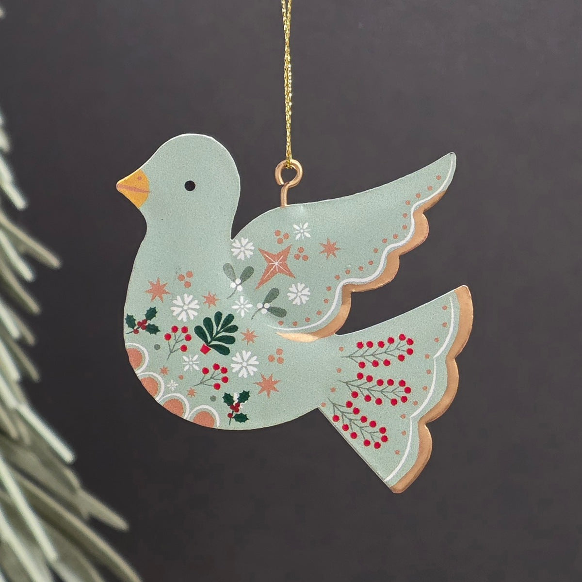 A decorative hanging ornament shaped like a dove, painted in a pale sage green with white flowers, red berries, green leaves, and a small orange beak. It hangs from a gold string against a dark grey background with blurred evergreen branches at the side.