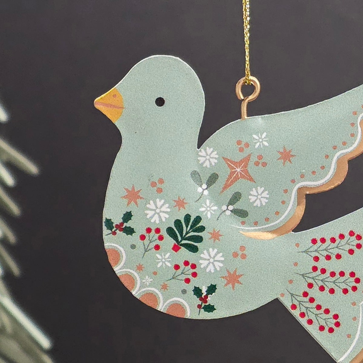 A decorative hanging ornament shaped like a dove, painted in a pale sage green with white flowers, red berries, green leaves, and a small orange beak. It hangs from a gold string against a dark grey background with blurred evergreen branches at the side.