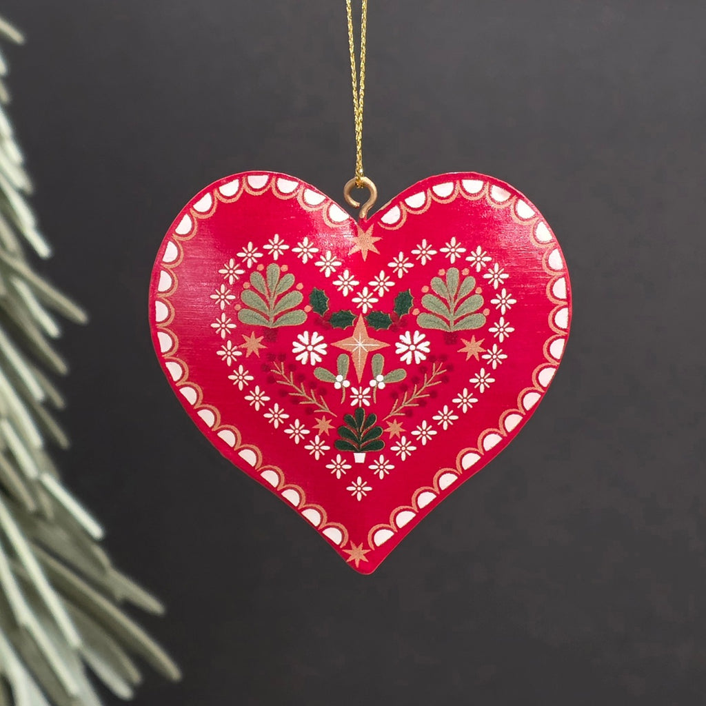 A red heart-shaped metal hanging ornament with hand-painted white, green, and gold floral and geometric patterns, hanging from a gold string against a dark background.